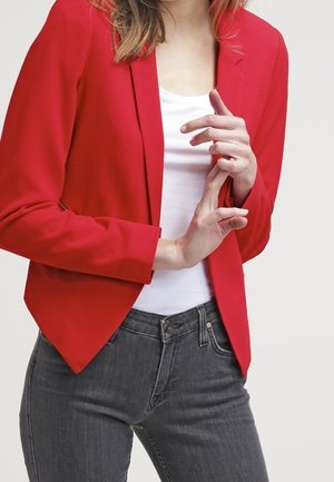 Vrouw draagt een rode blazer, wit shirt en donkere jeans, en stelt haar blazer met beide handen voor een effen achtergrond af.