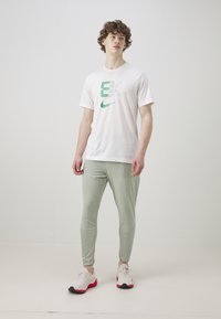 Hvit kortermet t-skjorte med grønt Nike-logo, parret med lysegrønne smale bukser og hvite-røde joggesko, stående mot en enkel bakgrunn.
