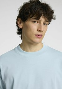 Šviesiai mėlynas apvalaus kaklo švarkelis, pagamintas iš minkštos medžiagos, su paprastu dizainu ir glotnia tekstūra, dėvimas žmogaus su trumpais, garbanotais plaukais.