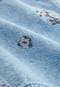 Lichtblauwe denim stof met kleine geborduurde witte en rode bloemen en groene bladeren in kruissteekstijl.