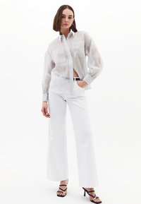 Durchsichtige weiße Bluse mit vertikalen grauen Streifen, zwei Brusttaschen, kombiniert mit hochgeschnittenen weißen Culottes und schwarzen Absätzen.
