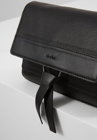 Sac en cuir noir avec rabat texturé et sangle lisse attachée à travers un anneau argenté, posé sur une surface blanche.