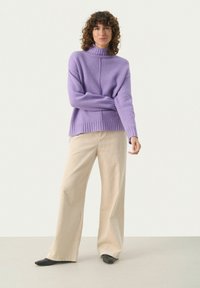Fioletowy sweter z golfem z dzianiny z ryflowanym dołem, w połączeniu z jasnobeżowymi szerokimi spodniami z korduroju i czarnymi butami typu slip-on.