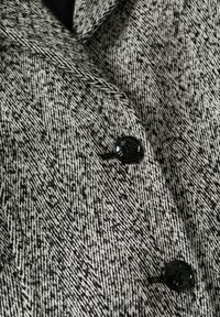 Grijs-zwart visgraatmotief stof met een gladde textuur, voorzien van twee glanzende zwarte knopen en reverskragdetails.