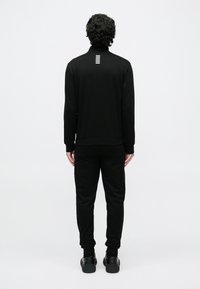 Mężczyzna z kręconymi włosami stoi tyłem, ubrany w czarny dres z małym białym logo w poziome paski na górnej części pleców.