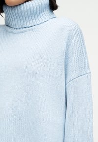 Femme portant un pull à col roulé en tricot bleu clair, montrant le haut du torse ainsi qu'une partie du cou et de l'épaule.