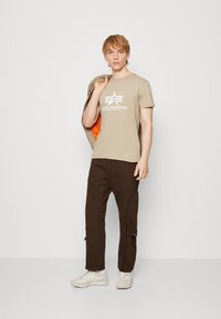 Tricou bej cu logo-ul "Alpha Industries", pantaloni cargo maro cu fermoare laterale și adidași de culoare deschisă. Modelul ține o jachetă.