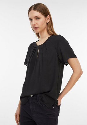 Frau mit glattem hellbraunem Haar, die eine lockere schwarze Kurzarmbluse und dunkle Hosen trägt und vor einem weißen Hintergrund steht.