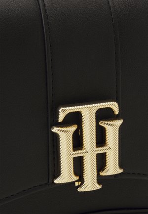Nærbillede af guld metalisk dobbelt T og H logo på sort struktureret læderoverflade med synlige syninger.