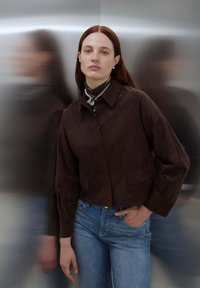 Braune Wildlederjacke in cropped Form mit Knopfverschluss, aufgesetzten Taschen und klassischem Kragen, kombiniert mit hellblauen Jeans.