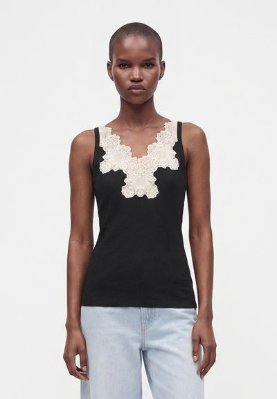 Woman wearing a black sleeveless top with wide beige lace detail at the neckline, paired with light blue jeans, standing against a plain background.