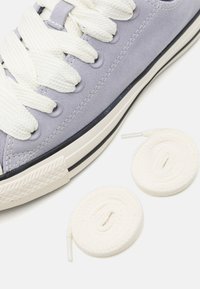 Sneaker viola in suede con lacci bianchi in cotone e suola in gomma. Due lacci bianchi arrotolati aggiuntivi sono posizionati accanto alla scarpa.