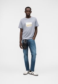 Ανοιχτό γκρι κοντομάνικη μπλούζα με ορθογώνιο γραφικό σχέδιο, συνδυασμένη με μπλε τζιν και μαύρη τσάντα χιαστί. Καθημερινά λευκά αθλητικά παπούτσια.