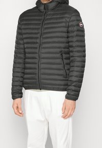 Man med en svart vadderad huva-jacka med framsidans dragkedja och dragkedjefickor, i kombination med vita byxor, mot en enfärgad bakgrund.