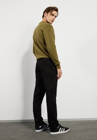 Súdadera de manga larga verde oliva, pantalones negros con bolsillos traseros y zapatillas negras con rayas blancas, vistas desde atrás sobre un fondo claro.