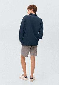 Tmavomodrá košeľa s dlhým rukávom na osobe, v kombinácii so svetlošedými šortkami a krémovými teniskami, z pohľadu zozadu. Hladká textúra látky.