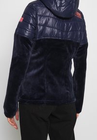 Henkilö, joka on pukeutunut tummansiniseen takkiin, jossa on tikkivuori yläselässä ja hupussa, sekä fleece-alaselkä ja hihat, yhdistettynä mustiin housuihin.