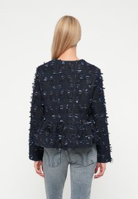 Jūras spēku peplum jaka, izgatavota no teksturēta auduma ar frēzēm un gaiši ziliem diegiem, ar garām piedurknēm un apaļu kakla izgriezumu.