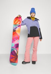 Snowboard s farebným abstraktným dizajnom, obsahujúcim ružové, modré a oranžové odtiene. Model má na sebe fialovo-čiernu bundu, ružové nohavice a modrý čiapku.
