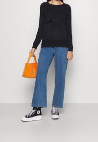 Czarny sweter ciążowy o fakturze z detalem w postaci kokardy, niebieskie spodnie typu wide-leg oraz pomarańczowa torebka z zamszu; zestawiony z czarno-białymi trampkami za kostkę.