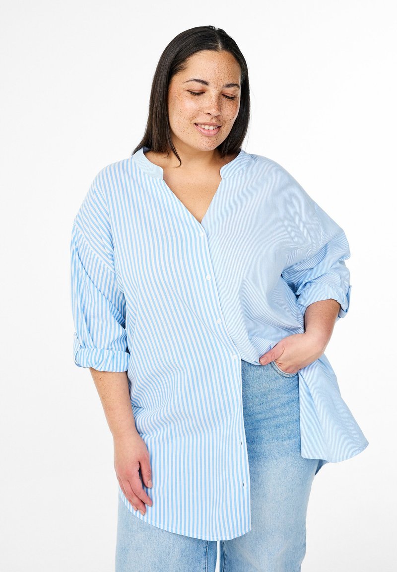 Camicetta azzurro chiaro con righe orizzontali e verticali, dotata di colletto alla cinese e maniche arrotolate, abbinata a denim chiaro.