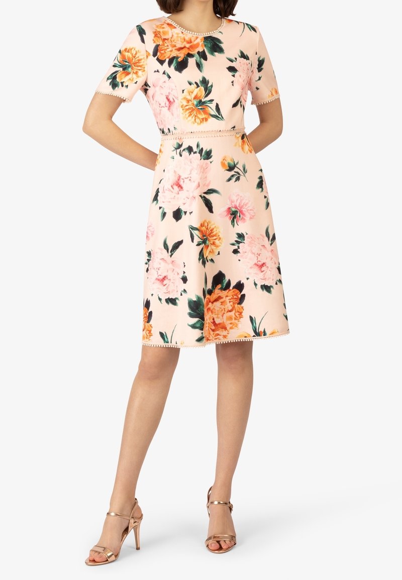 Femme portant une robe pêche courte à manches courtes avec un grand imprimé floral orange et rose et des sandales à talons hauts dorées.