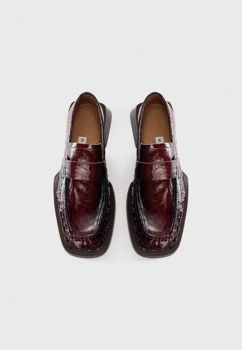 Une paire de mocassins en cuir rouge foncé brillant avec des orteils carrés et une semelle noire, vue de dessus sur un fond blanc.