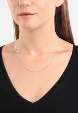 Donna che indossa una sottile collana d'argento sopra una maglietta nera a V, con lunghi capelli castani visibili su una spalla.