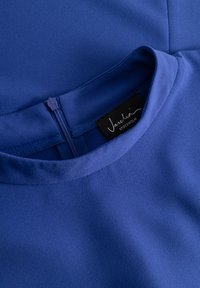 Blå blus med en mjuk textur, rund halsringning och dragkedja bak. Lappen säger "Vashti Stockholm." Materialet verkar lätt och flödande.