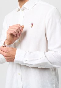 Valkoinen pellavapaita, jossa on napillinen kaulus, pitkät hihat ja pieni kirjailtu koriste rinnan kohdalla lähellä kaulusta.