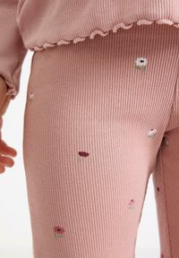 Lähikuva ribbaista vaaleanpunaisista housuista, joissa on pieniä brodeerattuja kukkia, joita lapsi pitää. Kuvassa näkyy myös osa vastaavasta yläosasta, jossa on röyhelöreuna.