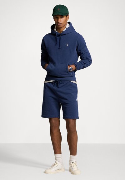 Felpa blu navy con tasca frontale e cordino, abbinata a pantaloni shorts blu navy con vita grigia chiara. Sneakers bianche e cappellino.