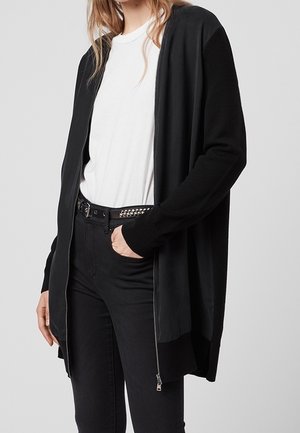 Femme portant une chemise blanche, un cardigan noir zippé, un pantalon noir et une ceinture noire cloutée, avec une main dans la poche.