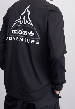 Černá košile s dlouhým rukávem s bílým logem Adidas Adventure a grafikou hory na zádech, kterou nosí osoba s jednou rukou v kapse.