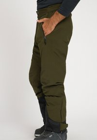 Pantaloni outdoor impermeabili in verde oliva con design tapered, tasche con zip, polsini regolabili e sezioni inferiori nere per maggiore resistenza.