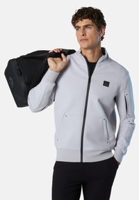 Felpa grigia chiara con zip in morbido tessuto, dotata di due tasche frontali, fessure laterali e un patch logo nero. Il modello tiene una borsa sportiva nera.