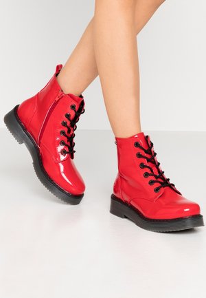 Nogi w błyszczących czerwonych sznurowanych botkach za kostkę z czarnymi podeszwami, bocznym zamkiem i pętelką na białym tle.