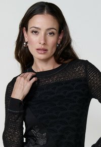Donna con lunghi capelli scuri, che indossa un maglione nero a fantasia e orecchini a cerchio d'argento, in posa con una mano sulla spalla.