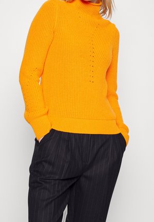 Persoon draagt een feloranje gebreide coltrui en zwarte broek met krijtstreep, handen in de zakken, op een effen witte achtergrond.