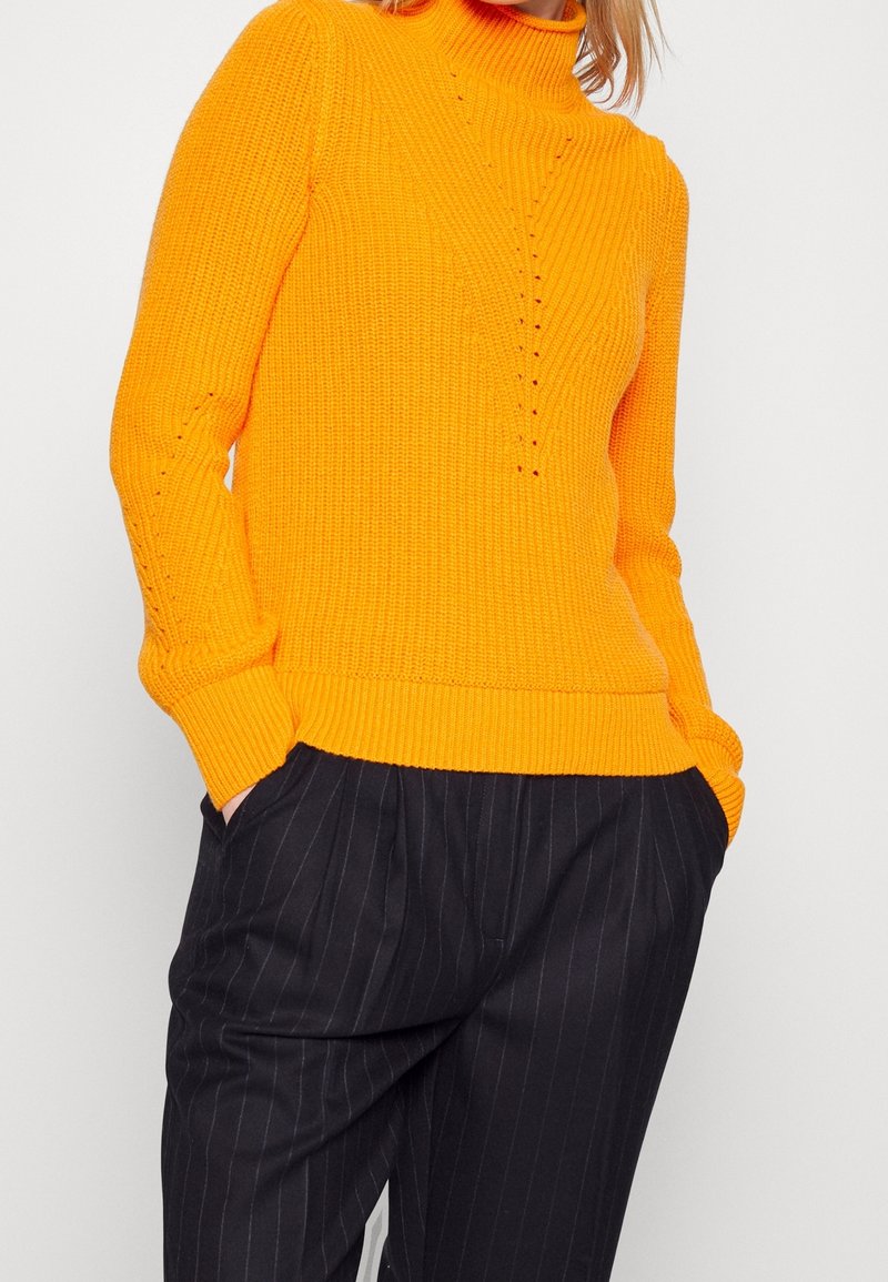 Personne portant un pull à col roulé en maille orange vif et un pantalon noir à rayures, les mains dans les poches, sur un fond blanc uni.