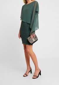 Haut en chiffon vert à couches associé à une jupe en dentelle verte. Les accessoires comprennent une pochette noire et dorée à motifs. Sandales à talons noires.