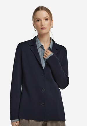 Marinefarbener Cardigan mit Reverskragen, langen Ärmeln und fünf vorderen Knöpfen. Hergestellt aus strukturiertem Stoff, über einem hellblau gestreiften Hemd getragen.
