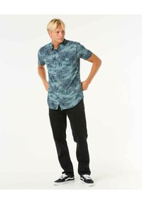 Korte mouwen, knoopshirt in teal met palmbomenprint, gecombineerd met zwarte broek en zwarte sneakers met een witte zool.