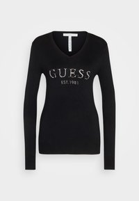 Zwarte v-hals gebreide trui met lange mouwen en een zilveren rhinestone logo "GUESS EST. 1981" over de borst. Zachte textuur.