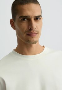 Camiseta de color claro, cuello redondo, hecha de material suave, con una textura lisa y un detalle de costura mínimo. Primer plano de la cara de un modelo masculino.