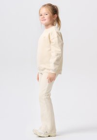 Jong meisje met licht haar in een paardenstaart, gekleed in een crèmekleurige sweatshirt, bijpassende broek en witte sneakers, staand in zijaanzicht op een witte achtergrond.