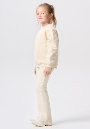 Junges Mädchen mit hellen Haaren im Pferdeschwanz, trägt einen cremefarbenen Pullover, passende Hose und weiße Turnschuhe, steht seitlich vor einem weißen Hintergrund.
