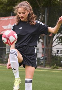 Juodas sportinis kostiumas su baltais dryžiais, iki kelių kojinės ir geltoni futbolo batai. Žaidėjas balansuoja ant raudono ir balto futbolo kamuolio.
