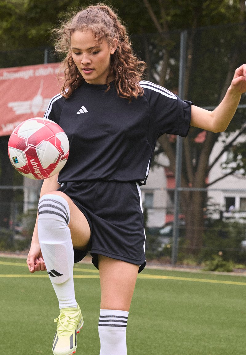 Juodas sportinis kostiumas su baltais dryžiais, iki kelių kojinės ir geltoni futbolo batai. Žaidėjas balansuoja ant raudono ir balto futbolo kamuolio.