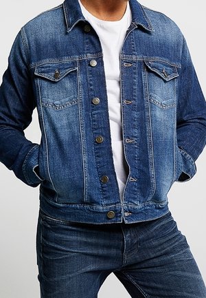Mann trägt eine blaue Jeansjacke über einem weißen Hemd, die Hände in den Jackentaschen, kombiniert mit blauen Jeans.